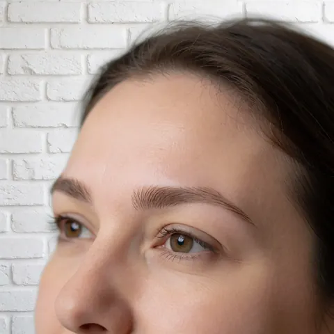 Close-up of a woman's face showing clean skin and well-defined eyebrows in front of a white brick wall, highlighting brow grooming details.