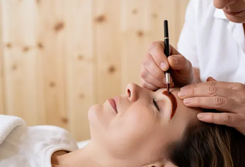 Professional applying henna to eyebrow with fine applicator against light wood wall during lamination service