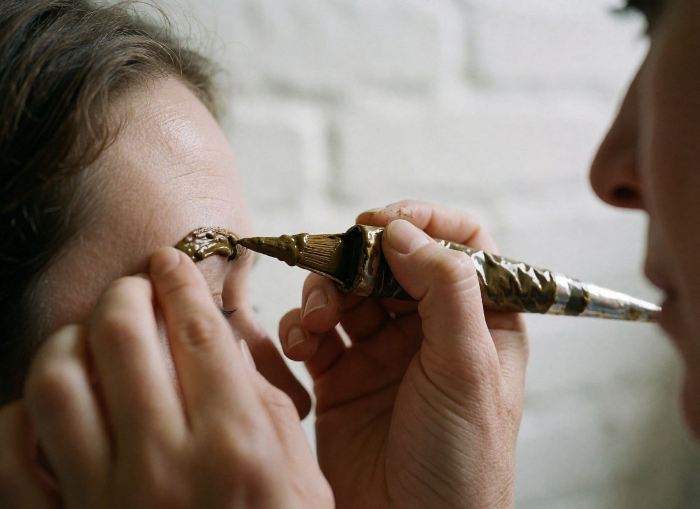 Close-up of a henna applicator touching skin near the eye during an eyebrow lamination service with a white brick wall background