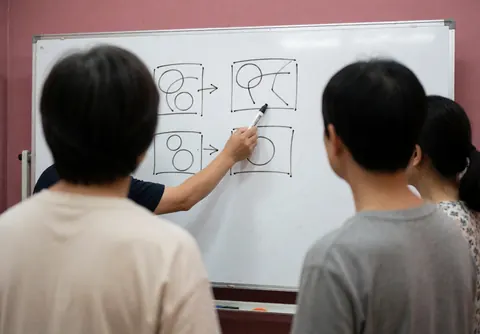 Instructor writing strategic diagrams on a whiteboard in front of students in a classroom with pink walls