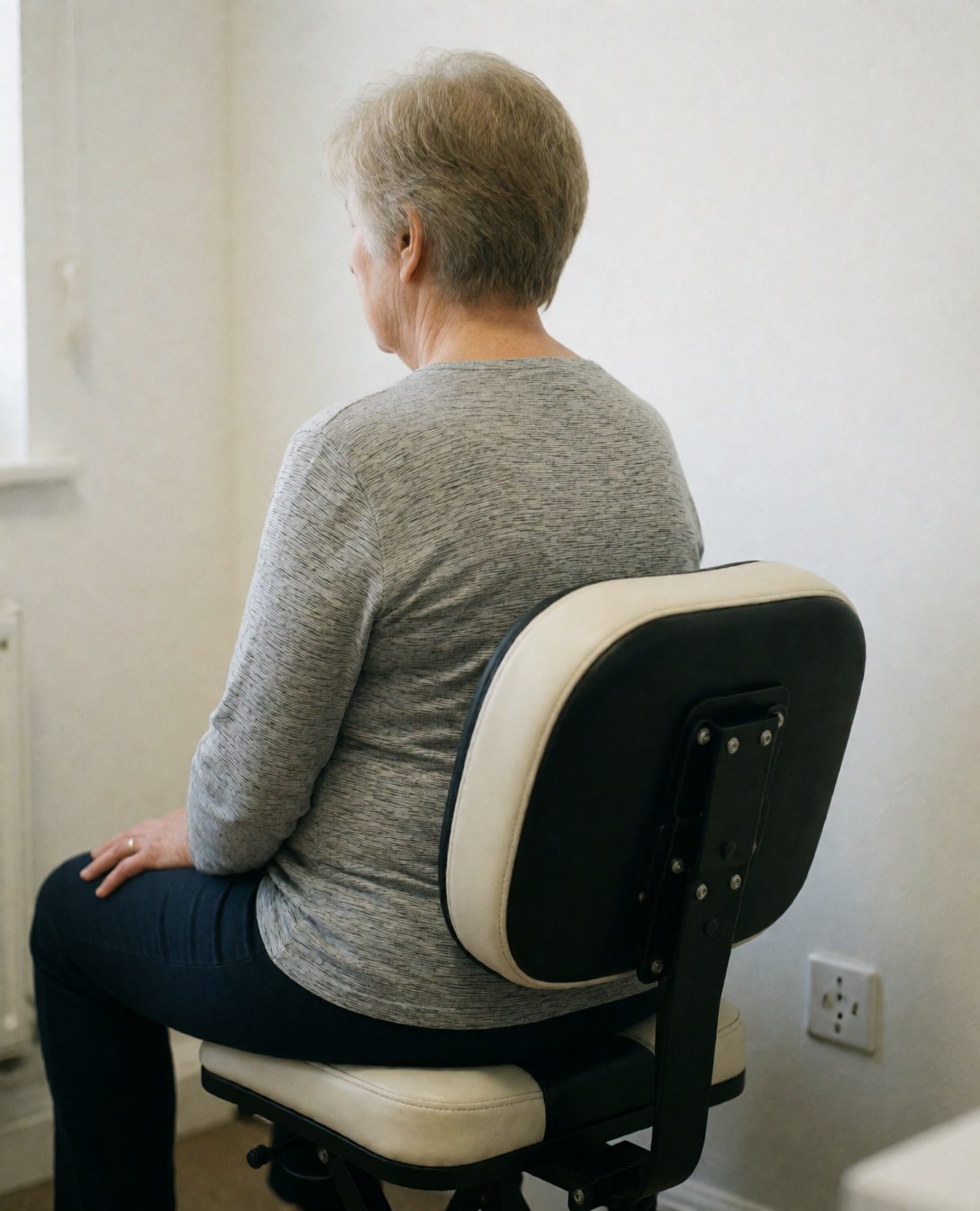 Senior woman seated in a massage chair featuring lumbar support, set against white walls with soft lighting in a geriatric care clinic.
