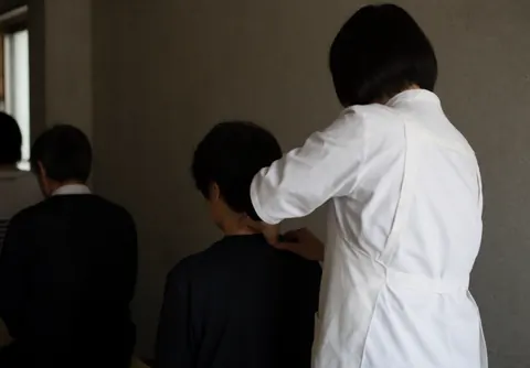 Thai massage practitioner performing a neck massage on a seated client in a calm setting with concrete walls and soft lighting.
