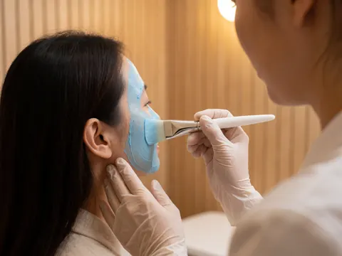 Aesthetician applies a blue hydrating face mask to a client's cheek using a white brush in a warm-lit beauty salon.