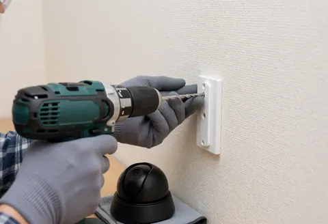 A technician wearing gloves uses a power drill to mount a security camera on a beige wall during an installation service.
