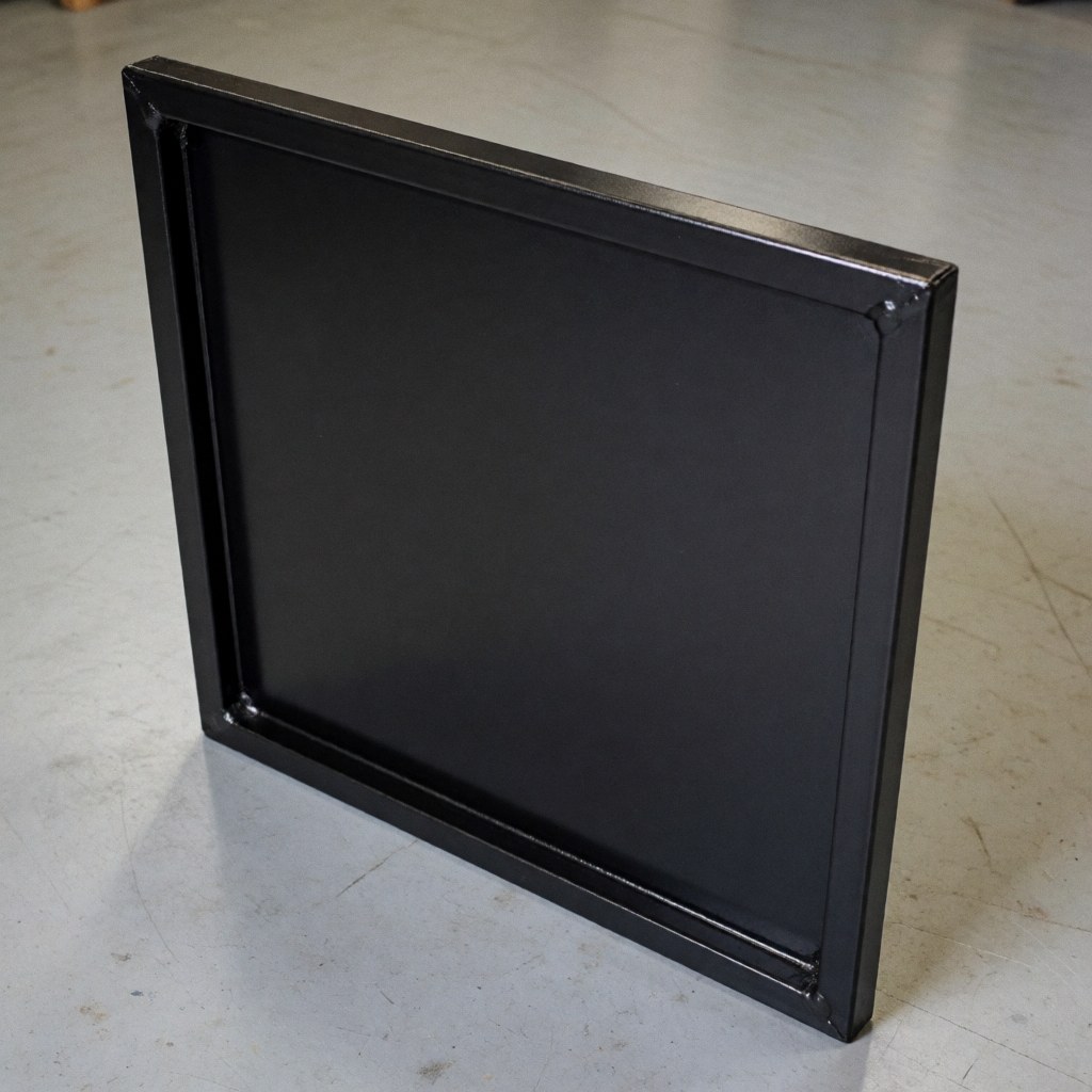 Close-up of a black matte painted metal repair panel showing welded seams on a concrete workshop floor.