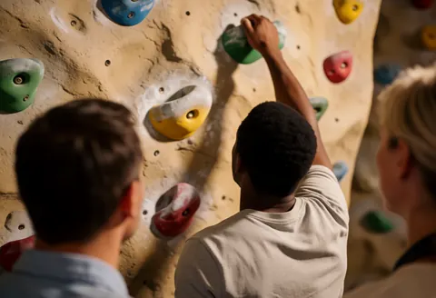 A rock climber reaches for a green handhold on an artificial climbing wall while others watch from the side.