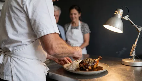 Close-up of a chef placing chicken with mole sauce onto white rice in a ceramic bowl under warm lighting in a kitchen setting.