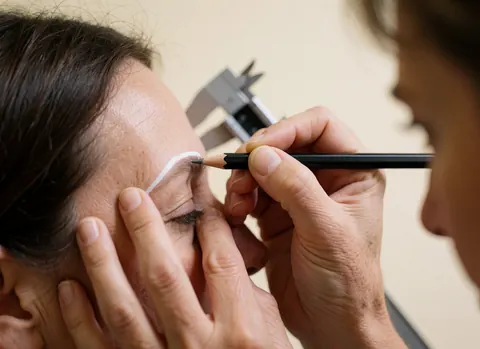 Close-up of a white pencil drawing a precise line on an eyebrow during a microblading service.