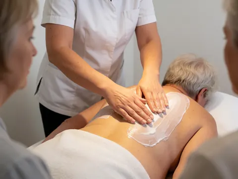 Close-up view of a therapist's hands applying white massage oil to a client's back during a professional session in a clean treatment room.