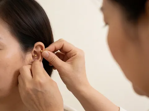 Close-up of hands placing golden drops into a patient's ear for auriculotherapy treatment with soft lighting and warm white walls.