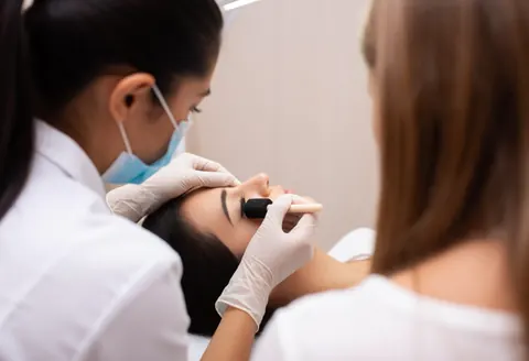 Professional lash technician wearing mask and gloves applies false eyelashes to a client using a foam brush in a salon setting.