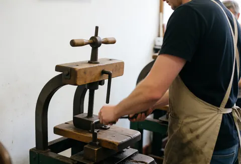 A cobbler working with a pressing tool in a workshop with a white wall and visible tools.