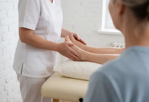 A therapist performs a lymphatic drainage massage on a client's foot in a treatment room with white brick walls and natural light.