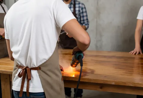 A technician wearing an apral uses a heat gun on a wooden table in a workshop with visible smoke and concrete walls.