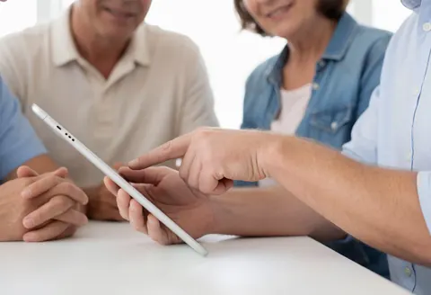 A professional smiles while showing a tablet to a client on a desk in a modern office setting.
