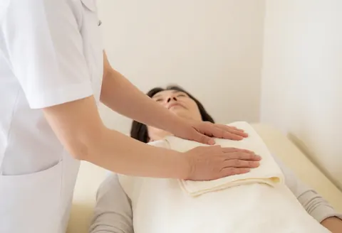 A therapist in a white uniform places a towel over a resting client's chest on a massage table.