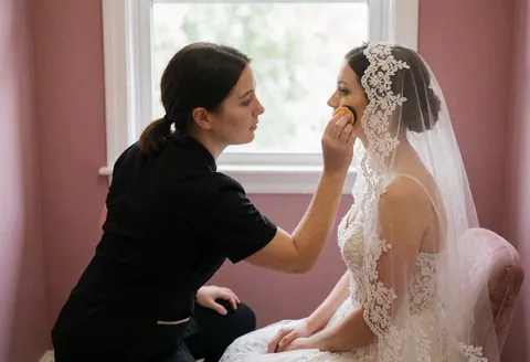 A professional makeup artist applies foundation to a bride wearing an embroidered veil in a room with pink walls and natural light.