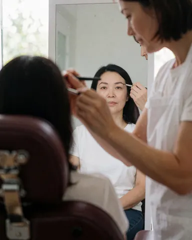 A technician applies makeup to a client's eyebrow while the client looks at her reflection in a mirror during a beauty salon service.
