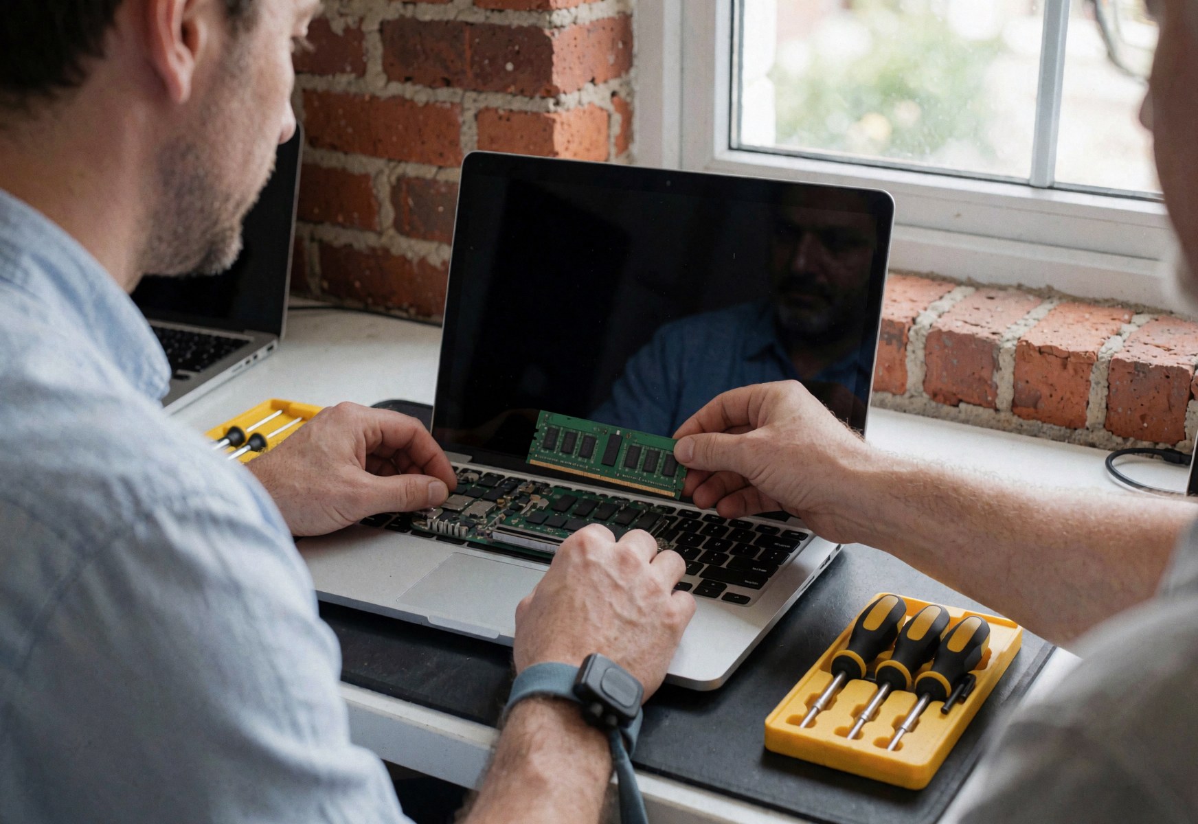 Close-up view of a technician installing green RAM into an open laptop on a workbench with tools and a brick wall background.