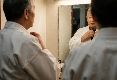 Person adjusting the collar of a white karate gi while looking into a mirror inside a dojo with warm white walls.