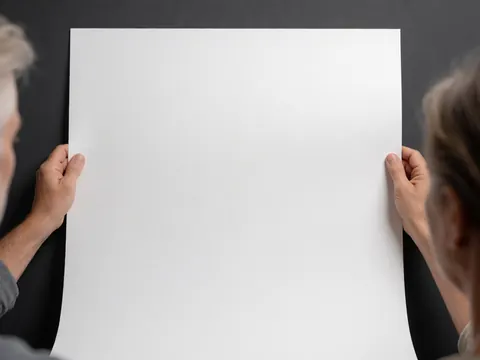 Hands holding a sheet of white paper against a gray wall, providing a clean background ideal for product presentation and graphic design layouts.