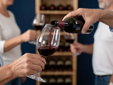 Red wine is being poured into a wine glass in front of a wooden shelf and dark blue wall, suitable for an online wine store.