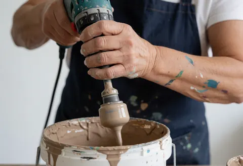 A hand uses an electric mixer to blend thick paint in a bucket during house painting service
