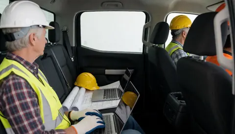 Two construction workers wearing hard hats and safety vests using laptops and blueprints inside a pickup truck for civil engineering project.