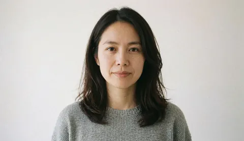 A professional woman wearing a gray sweater looks directly at the camera while standing in front of a clean white background for corporate team imagery.