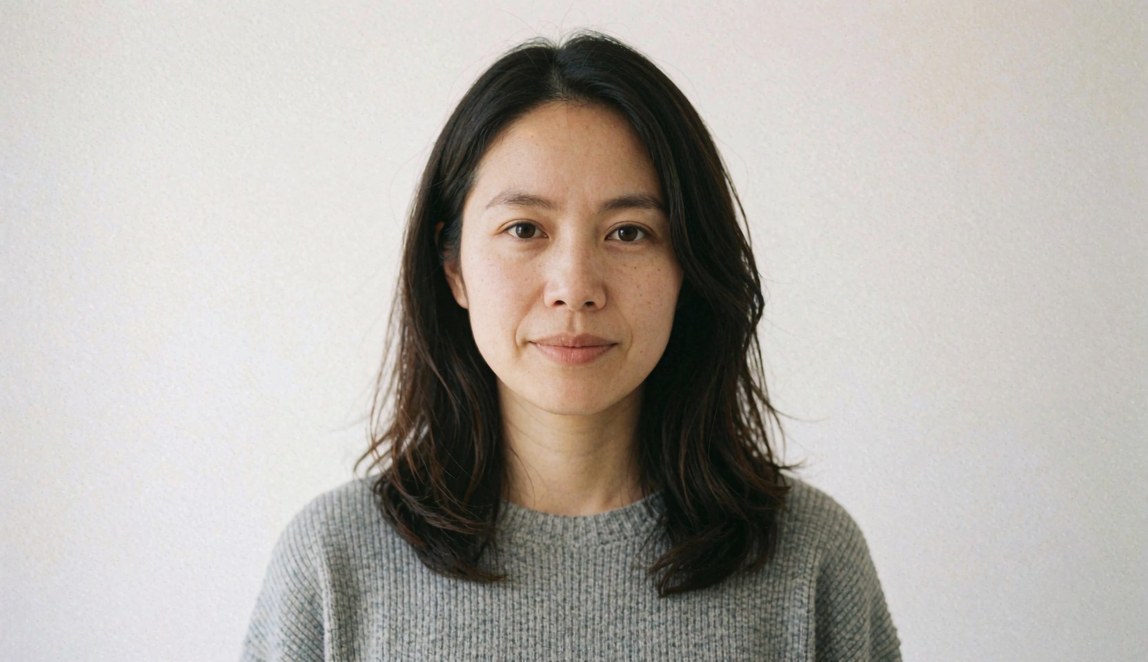 A professional woman wearing a gray sweater looks directly at the camera while standing in front of a clean white background for corporate team imagery.