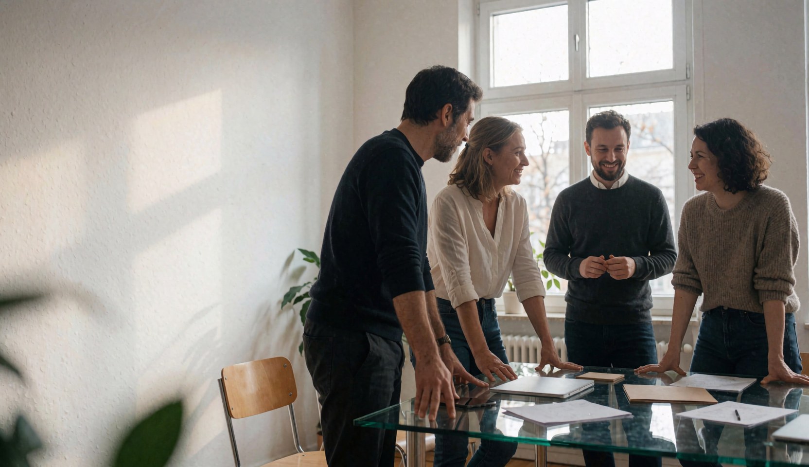 Four business professionals working together at a glass table in a bright modern office space with large windows and natural lighting.