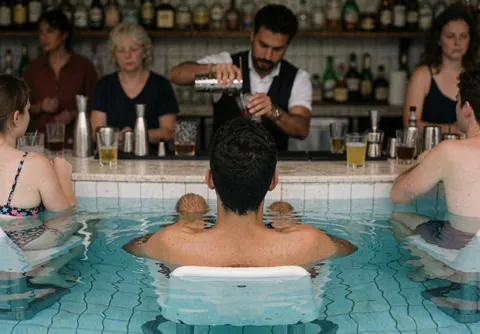 A bartender preparing beverages at a poolside bar with visible tiles and guests in the background.