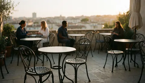 Empty hotel terrace featuring round wrought iron tables and chairs on stone paving with a city skyline visible in the background during sunset.