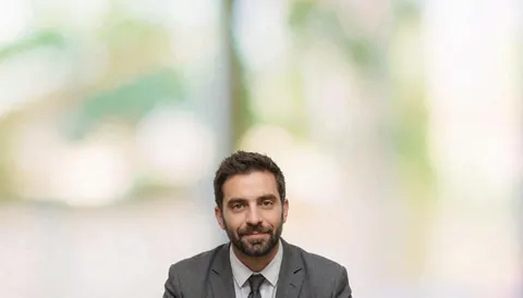 A businessman wearing a suit and tie looks directly at the camera against a blurred office background.
