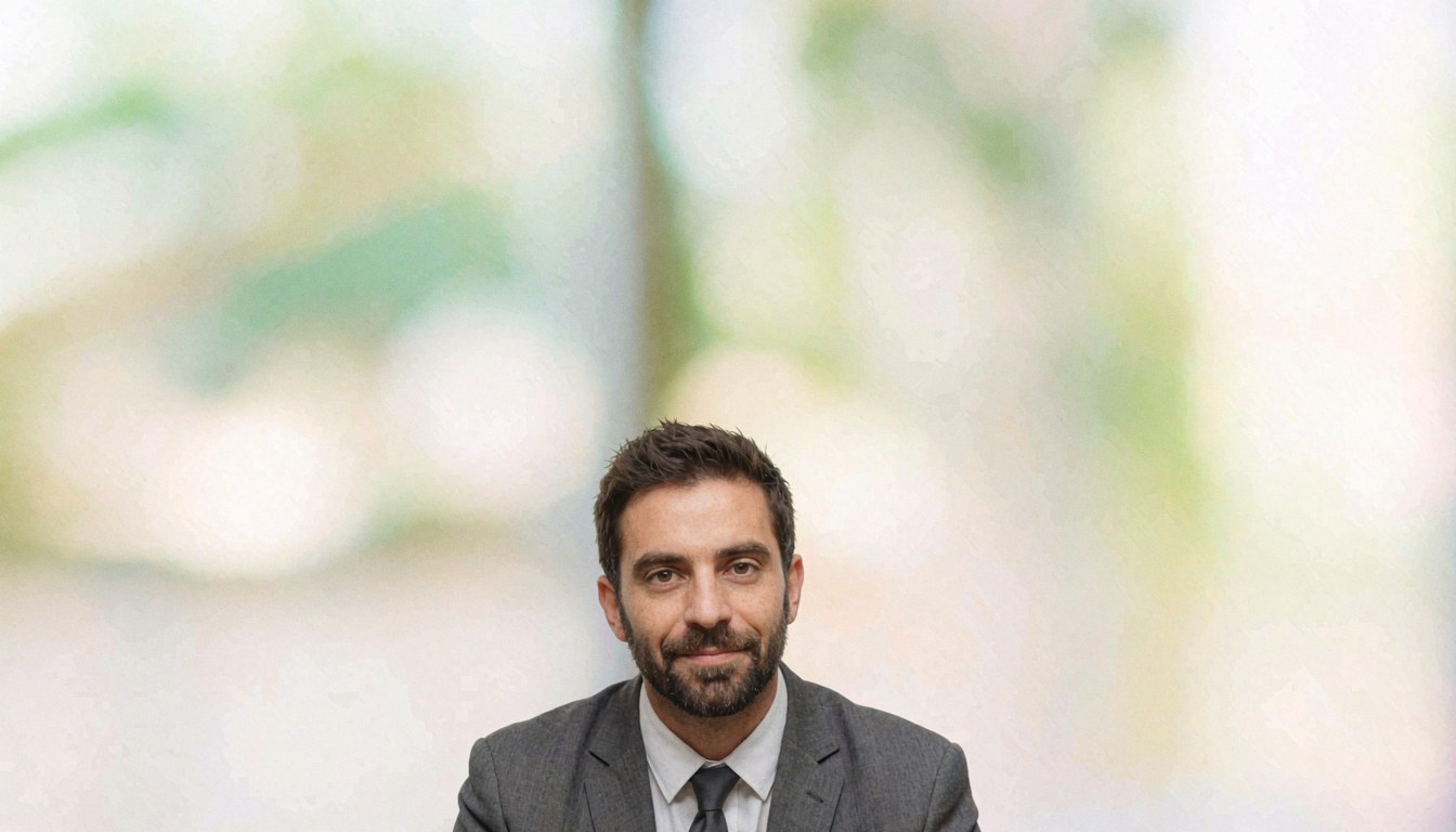 A businessman wearing a suit and tie looks directly at the camera against a blurred office background.