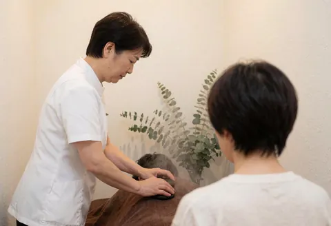 A therapist in white uniform applies hot stones to a client's back during a spa treatment with eucalyptus and warm white walls.