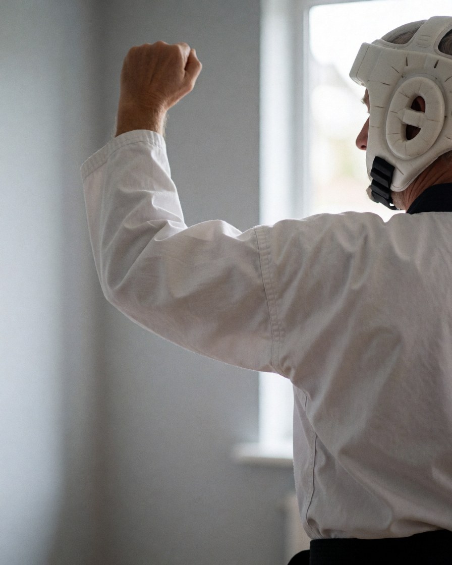 Martial arts instructor performing a block with a fist while wearing head protection and a white dobok in a room with light gray walls.