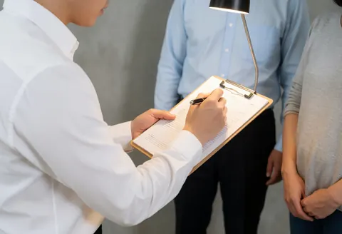 Insurance professional examines a document on a clipboard while seated at a concrete office desk under desk lamp light.