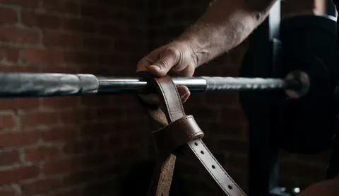 Close-up of a hand secured with leather straps around a weighted barbell in a brick gym setting for strength training.