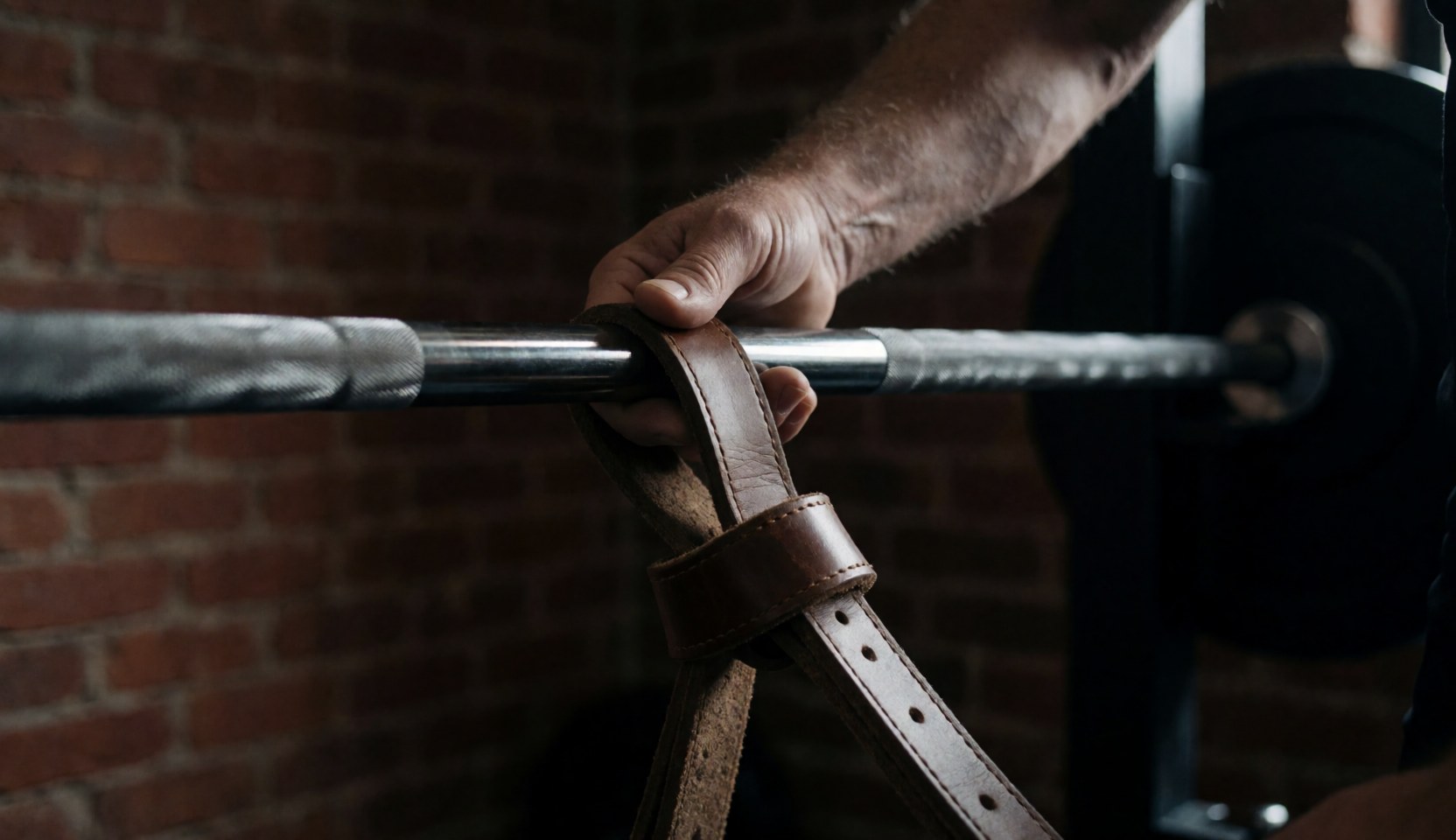 Close-up of a hand secured with leather straps around a weighted barbell in a brick gym setting for strength training.