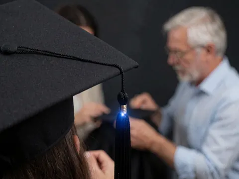 Close-up of a blue-lit tassel on a black graduation cap with hands adjusting it against a dark background.