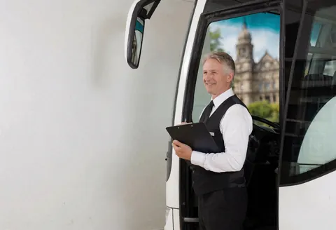 A bus driver in uniform holding a clipboard stands beside his vehicle in front of an old building.