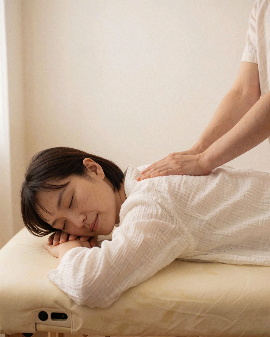 A Thai massage practitioner performs a treatment on a client lying face down on a white table in a calm setting.