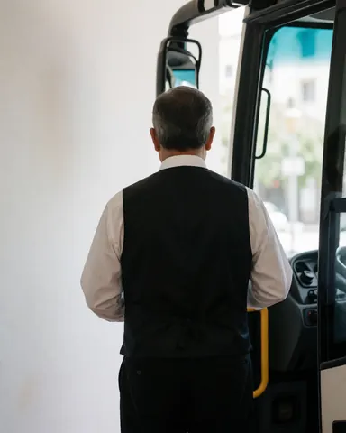 Back view of a tour bus driver in uniform standing next to the open door of the vehicle near a white wall at a hotel.