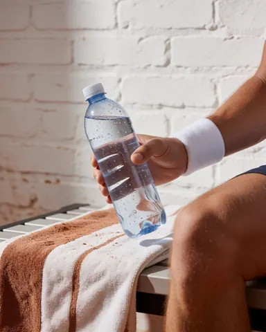 A hand holds a condensation-covered water bottle resting on a towel against a white brick background, suitable for tennis instructors.