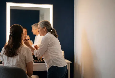 A makeup artist applies lipstick to a client seated before a mirror in a blue-walled studio with warm lighting.