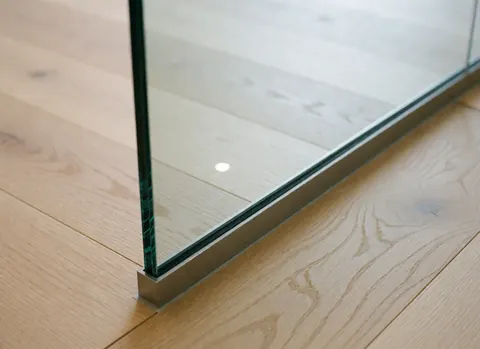 Close-up view of a frameless glass wall in a clinical setting with light wood flooring and stainless steel base reflecting ambient light.