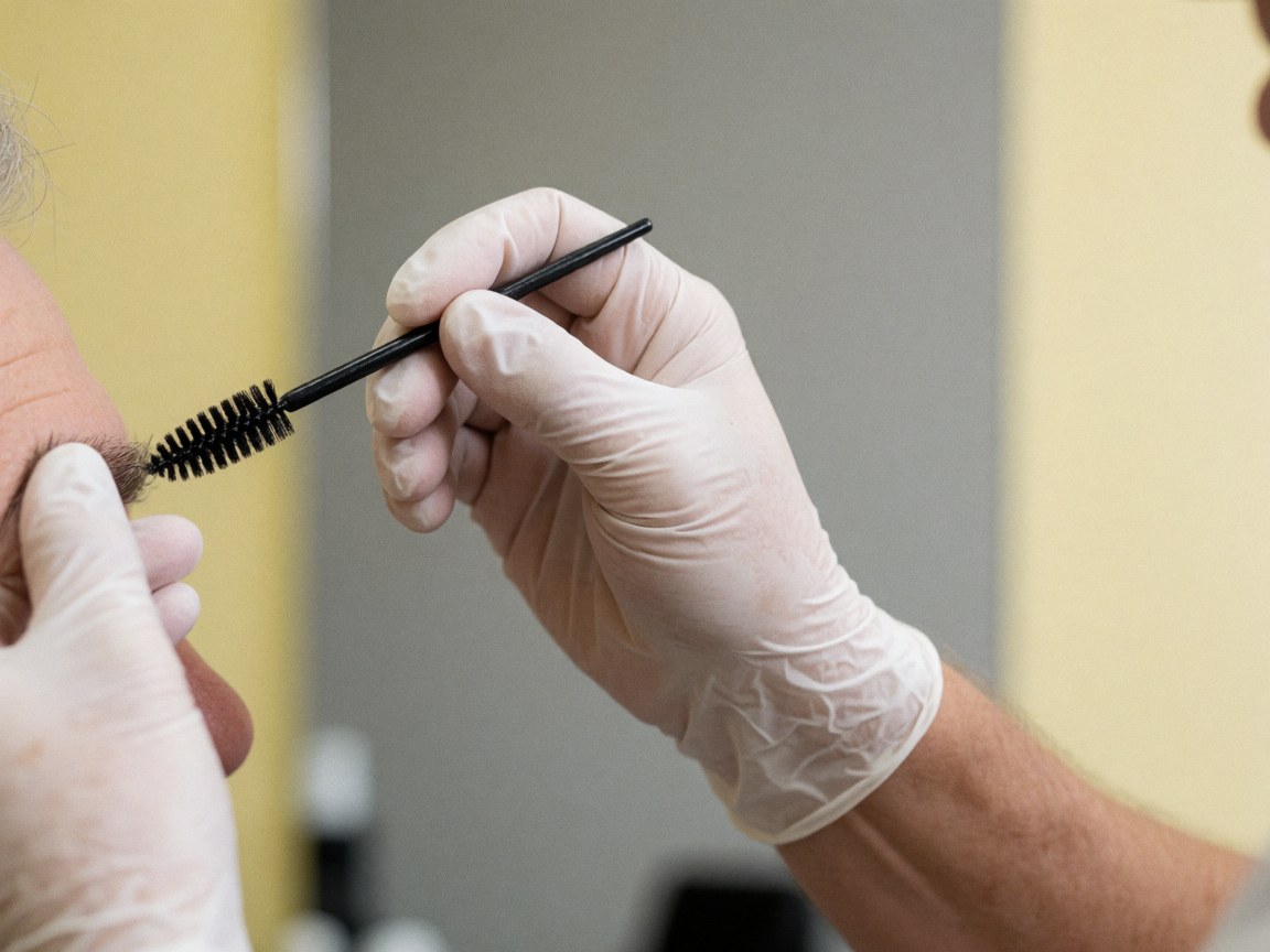 Close-up of a gloved hand using a brush to apply eyebrow product to a client in a barbershop with yellow walls and a neutral gray background.
