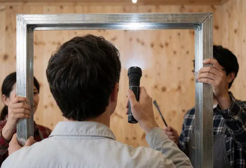 Three workers use a flashlight to inspect a metal frame in a workshop with wooden walls.