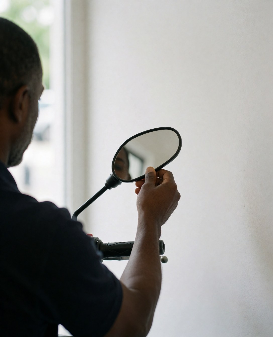 A motorbike taxi driver adjusts the side mirror of his vehicle against a plain white wall in natural daylight.